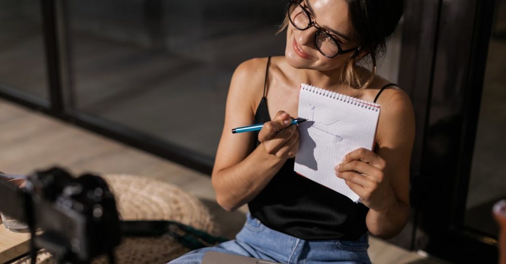 Smiling woman vlogging indoors with a camera, laptop, and notepad.
